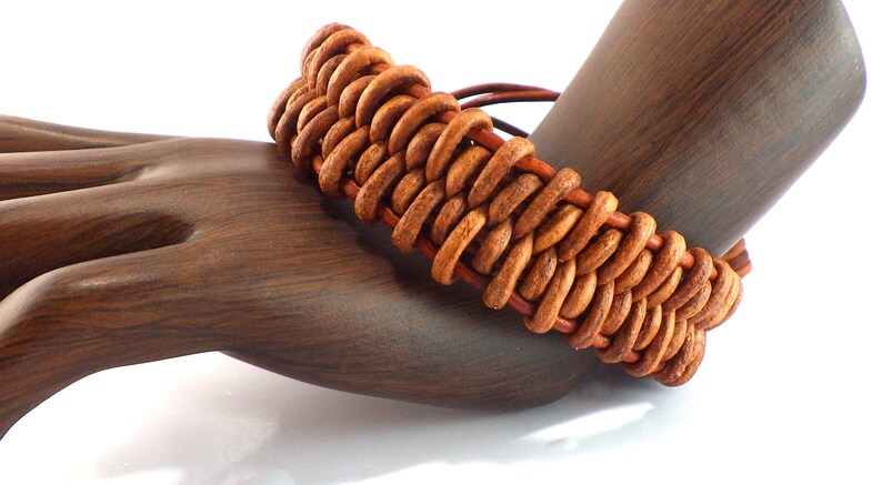 Puede incluir: Una pulsera de cuero marr&oacute;n hecha a mano con un dise&ntilde;o trenzado. La pulsera se exhibe en un soporte para joyas con forma de mano de madera, mostrando sus intrincados detalles y tonos c&aacute;lidos. El dise&ntilde;o de la pulsera presenta una serie de bucles de cuero interconectados.