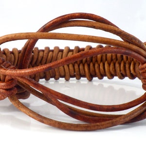 Puede incluir: Pulsera de cuero marr&oacute;n con una secci&oacute;n central trenzada y nudos decorativos. La pulsera est&aacute; hecha de m&uacute;ltiples cordones de cuero, creando un accesorio texturizado y elegante. El dise&ntilde;o incorpora una secci&oacute;n central de hebras de cuero muy tejidas.