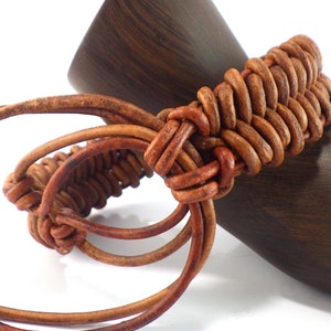 Puede incluir: Un brazalete de cuero marr&oacute;n con un dise&ntilde;o tejido. El brazalete est&aacute; hecho de bucles de cuero entrelazados. El brazalete se muestra sobre un fondo de madera oscura. El brazalete es de color marr&oacute;n c&aacute;lido.