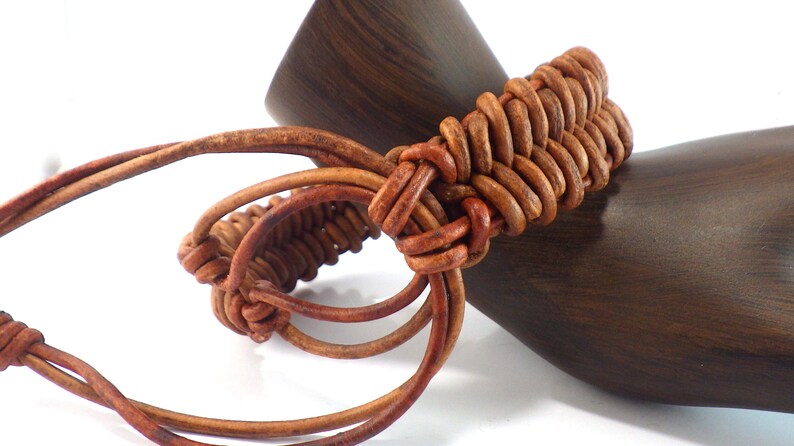 Puede incluir: Un brazalete de cuero marr&oacute;n con un dise&ntilde;o tejido. El brazalete est&aacute; hecho de bucles de cuero entrelazados. El brazalete se muestra sobre un fondo de madera oscura. El brazalete es de color marr&oacute;n c&aacute;lido.