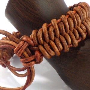 Puede incluir: Una pulsera de cuero marr&oacute;n con un dise&ntilde;o tejido. La pulsera est&aacute; hecha de m&uacute;ltiples hebras de cuero, intrincadamente anudadas. La pulsera se muestra en una forma de madera marr&oacute;n oscuro.