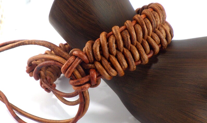 Puede incluir: Una pulsera de cuero marr&oacute;n con un dise&ntilde;o tejido. La pulsera est&aacute; hecha de m&uacute;ltiples hebras de cuero, intrincadamente anudadas. La pulsera se muestra en una forma de madera marr&oacute;n oscuro.