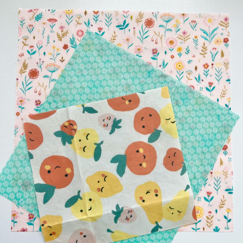 May include: A collection of reusable food wraps with colorful patterns. One wrap features a light pink floral design, another has a teal floral pattern, and the third displays smiling oranges, lemons, and pears on a white background. These wraps are an eco-friendly alternative to plastic.