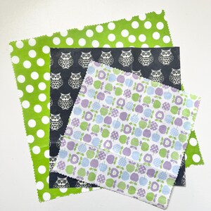 May include: A collection of reusable food wraps in various patterns. One wrap is green with white polka dots, another is dark gray with white owl illustrations, and the third is white with green, blue, and purple apple designs. These wraps are a sustainable alternative to plastic.