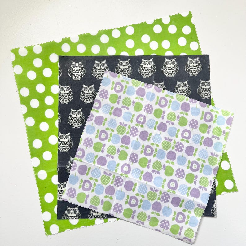 May include: A collection of reusable food wraps in various patterns. One wrap is green with white polka dots, another is dark gray with white owl illustrations, and the third is white with green, blue, and purple apple designs. These wraps are a sustainable alternative to plastic.