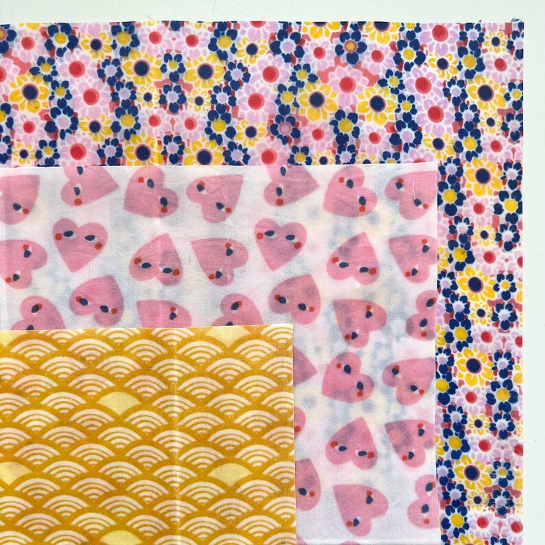 May include: A collection of patterned fabric squares. The top fabric features a pink floral design with yellow and blue accents. Below, a white fabric with pink heart shapes is visible. The bottom fabric has a yellow, wave-like pattern.