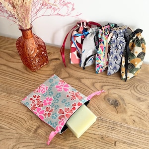 May include: A collection of small fabric drawstring pouches featuring various colorful patterns, including floral, geometric, and fan-shaped designs. One pouch in the foreground is light gray with pink and teal flowers, containing a rectangular soap bar.