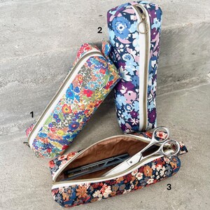 May include: Three floral print pencil cases in various colors. One case is open, revealing scissors and pens. The cases have white zippers and are made of a colorful floral fabric. The background is a neutral gray surface.
