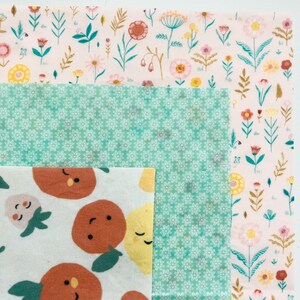 May include: A collection of reusable food wraps in various floral and fruit patterns. The wraps feature a light pink background with colorful flowers, a turquoise background with white flowers, and a white background with orange and yellow fruit.