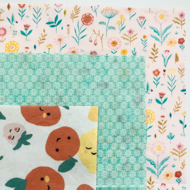 May include: A collection of reusable food wraps in various floral and fruit patterns. The wraps feature a light pink background with colorful flowers, a turquoise background with white flowers, and a white background with orange and yellow fruit.