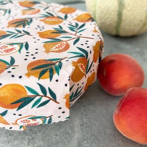 May include: A reusable food wrap featuring a light pink background with a repeating pattern of orange pomegranates, green leaves, and small black dots. The fabric wrap is shown covering the top of a round container, displaying a folded edge.