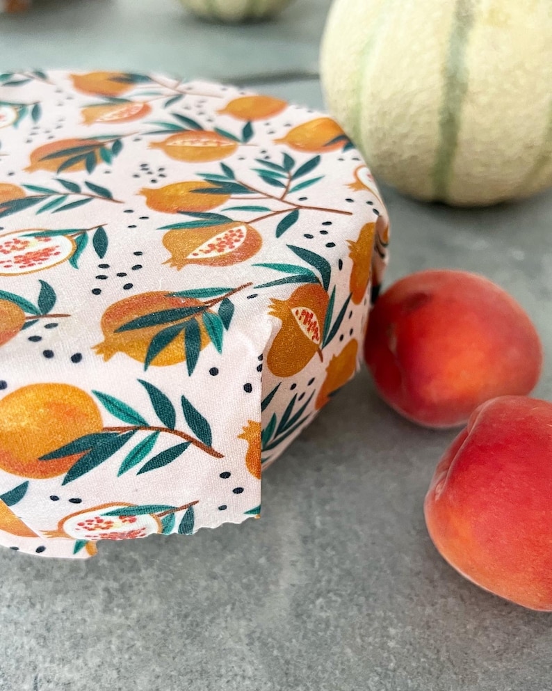 May include: A reusable food wrap featuring a light pink background with a repeating pattern of orange pomegranates, green leaves, and small black dots. The fabric wrap is shown covering the top of a round container, displaying a folded edge.