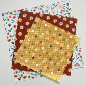 May include: Three reusable beeswax food wraps with floral patterns. One wrap is light blue with pink and yellow flowers, another is brown with white daisies, and the third is yellow with a gingham pattern and small flowers.