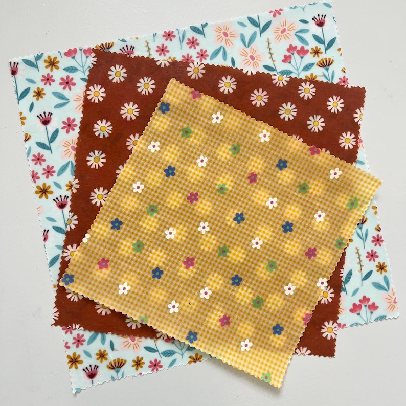 May include: Three reusable beeswax food wraps with floral patterns. One wrap is light blue with pink and yellow flowers, another is brown with white daisies, and the third is yellow with a gingham pattern and small flowers.