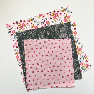 May include: Three reusable beeswax food wraps with floral and botanical patterns. The top wrap has a white background with pink and red flowers. The middle wrap has a dark green background with a tropical leaf design. The bottom wrap is pink with small red flowers.