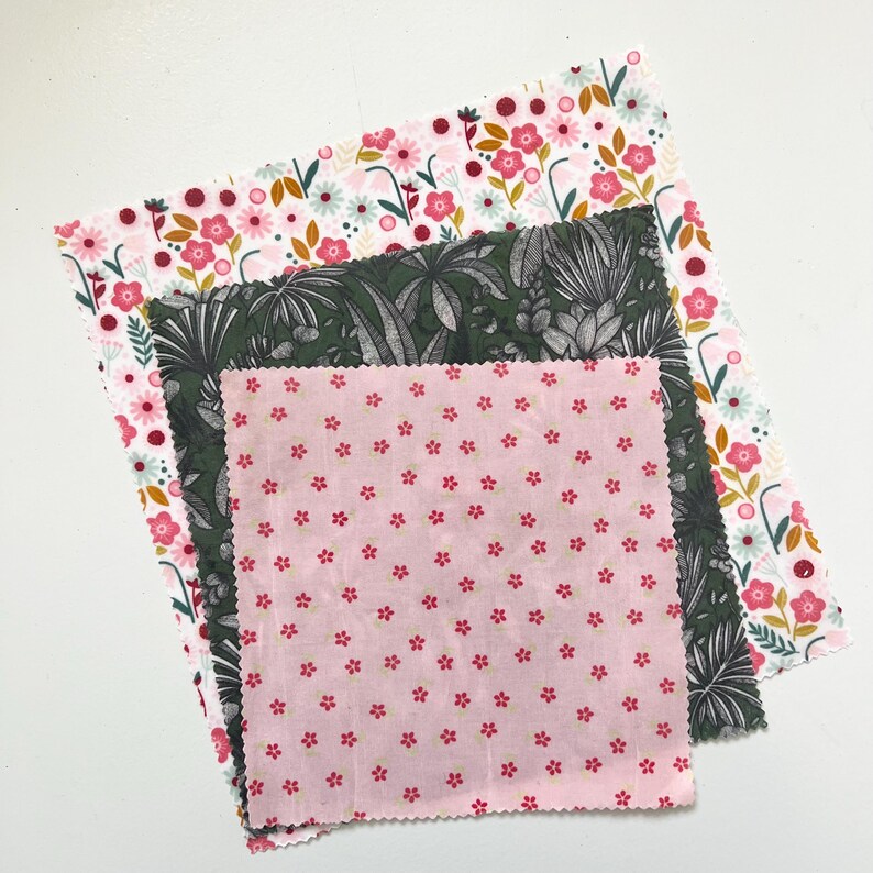 May include: Three reusable beeswax food wraps with floral and botanical patterns. The top wrap has a white background with pink and red flowers. The middle wrap has a dark green background with a tropical leaf design. The bottom wrap is pink with small red flowers.
