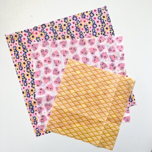 May include: Three square fabric wraps featuring distinct patterns. The bottom layer has a dark blue background with yellow and pink flowers. The middle layer is white with pink heart shapes. The top layer is yellow with a white wave pattern.