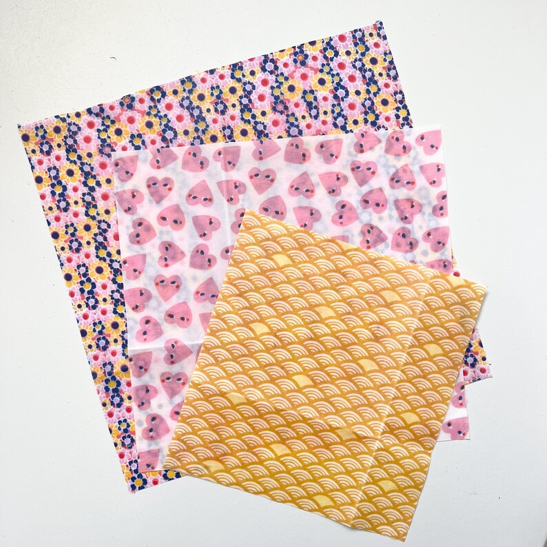May include: Three square fabric wraps featuring distinct patterns. The bottom layer has a dark blue background with yellow and pink flowers. The middle layer is white with pink heart shapes. The top layer is yellow with a white wave pattern.