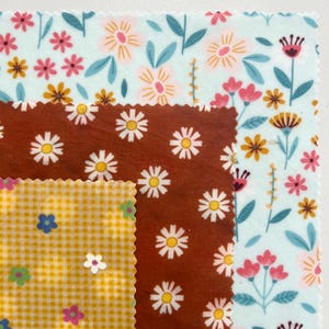 May include: Three stacked square fabric wraps with pinked edges. The bottom layer is light blue with a multi-colored floral pattern. The middle layer is rust brown with a white daisy print. The top layer is yellow gingham with small flowers.