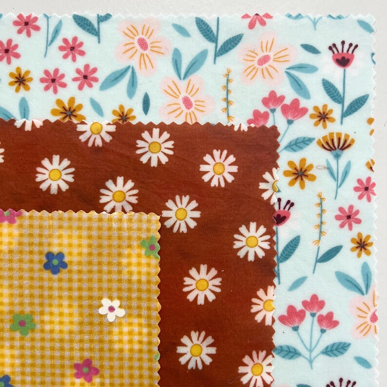 May include: Three stacked square fabric wraps with pinked edges. The bottom layer is light blue with a multi-colored floral pattern. The middle layer is rust brown with a white daisy print. The top layer is yellow gingham with small flowers.