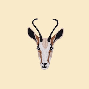 Può includere: Toppa ricamata raffigurante la testa di un'antilope saltante. La toppa ha uno sfondo color crema con una rappresentazione dettagliata del volto dell'animale, inclusi accenti neri e marroni sulle orecchie e sulle corna. Il volto dell'antilope saltante è bianco con dettagli marroni.