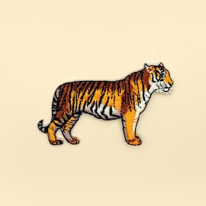 Può includere: Una toppa ricamata da stirare con un tigre arancione e nero in piedi su uno sfondo beige.