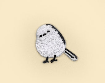 May include: A white and black embroidered patch of a long-tailed tit bird with a black head and a white body.