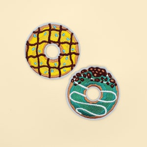 Peut inclure: Deux patchs de beignets brodés. Un beignet est jaune avec un glaçage brun et des pépites bleues. L'autre beignet est vert avec des pépites brunes et un glaçage blanc.