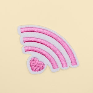 Puede incluir: Parche de arcoíris bordado en rosa con un pequeño corazón rosa en el centro.