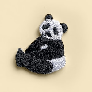 Pode incluir: Patch bordado de panda em preto, branco e cinza. O panda está sentado com os braços e as pernas dobrados. O patch é feito de fio e tem um design detalhado.