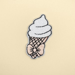May include: White embroidered ice cream cone with a pink bow. The ice cream is in a waffle cone.