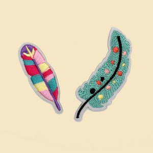 May include: Two embroidered feather patches. One is a colorful design with pink, red, teal, and yellow. The other is teal with red, yellow, and black accents. Both have a gray border.