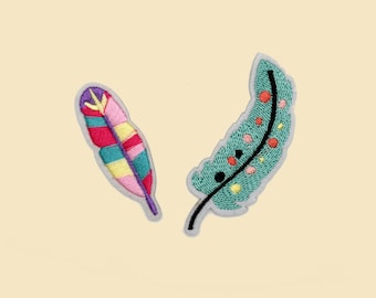Könnte beinhalten: Zwei gestickte Federaufnäher. Einer ist ein farbenfrohes Design mit Rosa, Rot, Türkis und Gelb. Der andere ist türkis mit roten, gelben und schwarzen Akzenten. Beide haben einen grauen Rand.