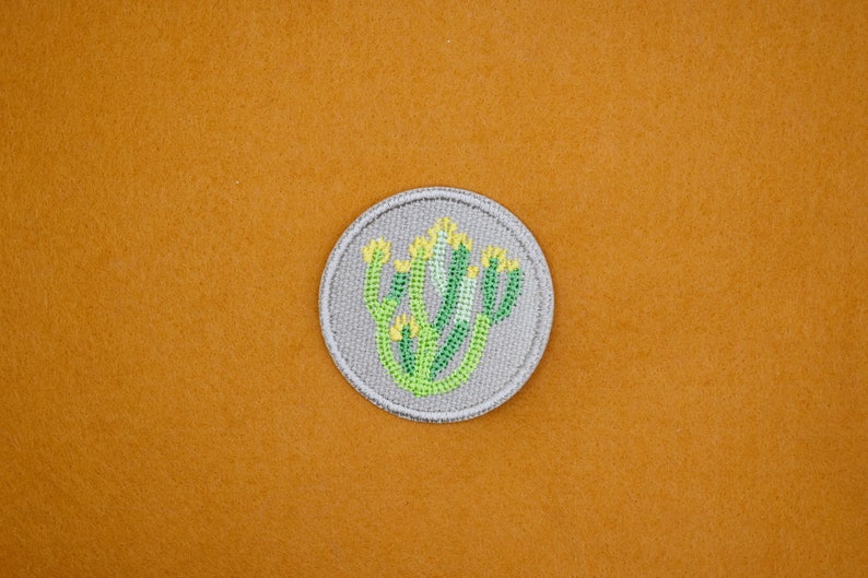 May include: A round, grey embroidered patch featuring a green and yellow cactus design.