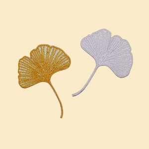 May include: Two embroidered ginkgo leaf patches. One is gold, the other is silver. The leaves have detailed stitching and are set against a light beige background. These patches could be used for clothing or craft projects.