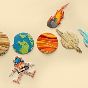 May include: A set of embroidered iron-on patches featuring planets, a comet, and an astronaut. The planets include a brown and orange Jupiter, a blue and green Earth, a red and orange Mars, a beige Saturn with rings, and a blue Neptune with rings. The comet is orange and yellow with a grey rock. The astronaut is wearing a white spacesuit with an orange visor and is floating in space.