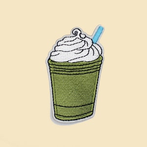 May include: Green and white embroidered patch of a green iced coffee drink with whipped cream and a blue straw.