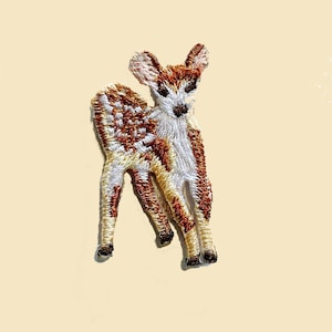 Puede incluir: Parche bordado para planchar de un cervatillo marrón y blanco con manchas blancas. El cervatillo está de pie sobre sus cuatro patas con la cabeza girada hacia un lado.
