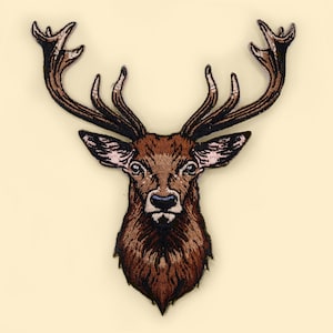 以下が含まれることがあります： 大きな角を持つ茶色の鹿の頭が刺繍されたパッチ。鹿は濃い茶色の鼻と大きく暗い目をしています。
