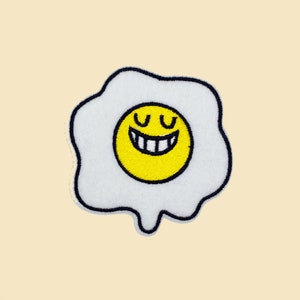 May include: A white embroidered patch shaped like a fried egg with a yellow smiling face in the center.