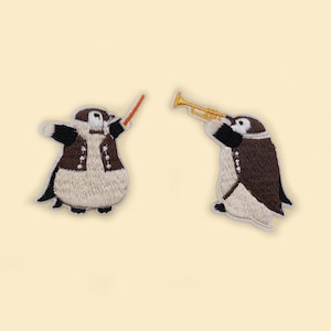 Può includere: Due toppe ricamate di pinguini, uno suona la tromba e l'altro dirige con una bacchetta. I pinguini indossano giacche marroni e hanno la pancia bianca.