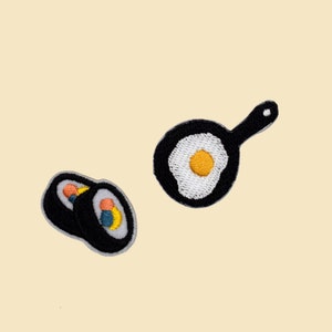 May include: Two embroidered iron-on patches, one of a black frying pan with a white egg inside and the other of two black sushi rolls with colorful fillings.