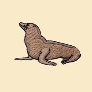 Puede incluir: Parche bordado con un sello marrón en posición sentada, mirando hacia arriba. El sello tiene costuras detalladas y está sobre un fondo beige claro. Ideal para chaquetas, bolsos u otros artículos de tela.