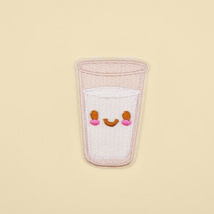 May include: A white embroidered patch depicting a glass of milk with a smiling face. The glass is pink and the face has brown eyes and a pink mouth.