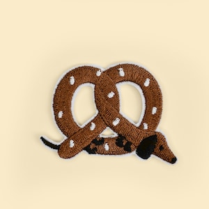May include: Embroidered patch featuring a brown pretzel design with white polka dots and a dachshund dog. The patch has a white border and is set against a light yellow background.