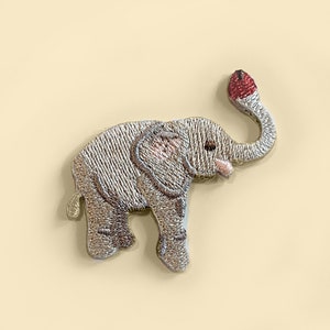 May include: A small, embroidered elephant patch with a grey body, pink ears, and a red tip on its trunk.