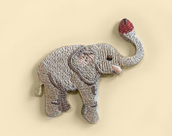 May include: A small, embroidered elephant patch with a gray body, pink ears, and a red tip on its trunk.