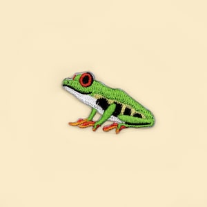 May include: Green and white embroidered frog patch with red eyes. The frog is sitting on a pale yellow background.