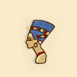 May include: Embroidered patch featuring the profile of Nefertiti, the Great Royal Wife of the Egyptian Pharaoh Akhenaten. The patch has a blue headdress with red and yellow accents, a tan face, and a brown border.