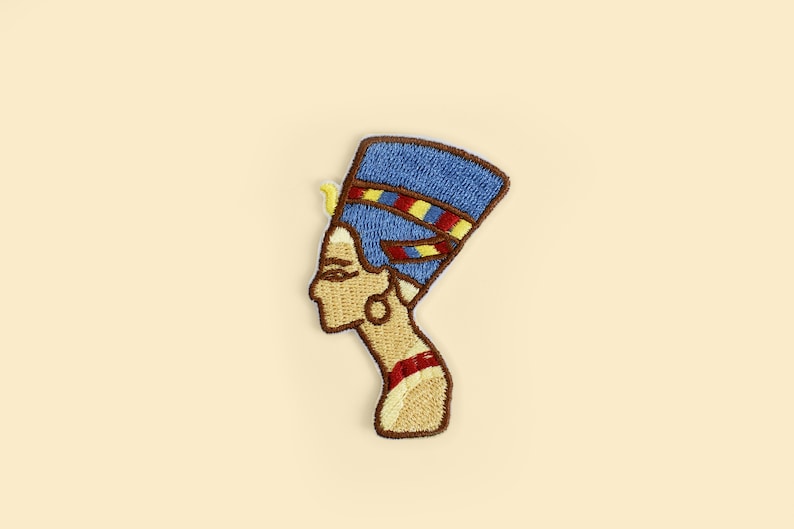 May include: Embroidered patch featuring the profile of Nefertiti, the Great Royal Wife of the Egyptian Pharaoh Akhenaten. The patch has a blue headdress with red and yellow accents, a tan face, and a brown border.